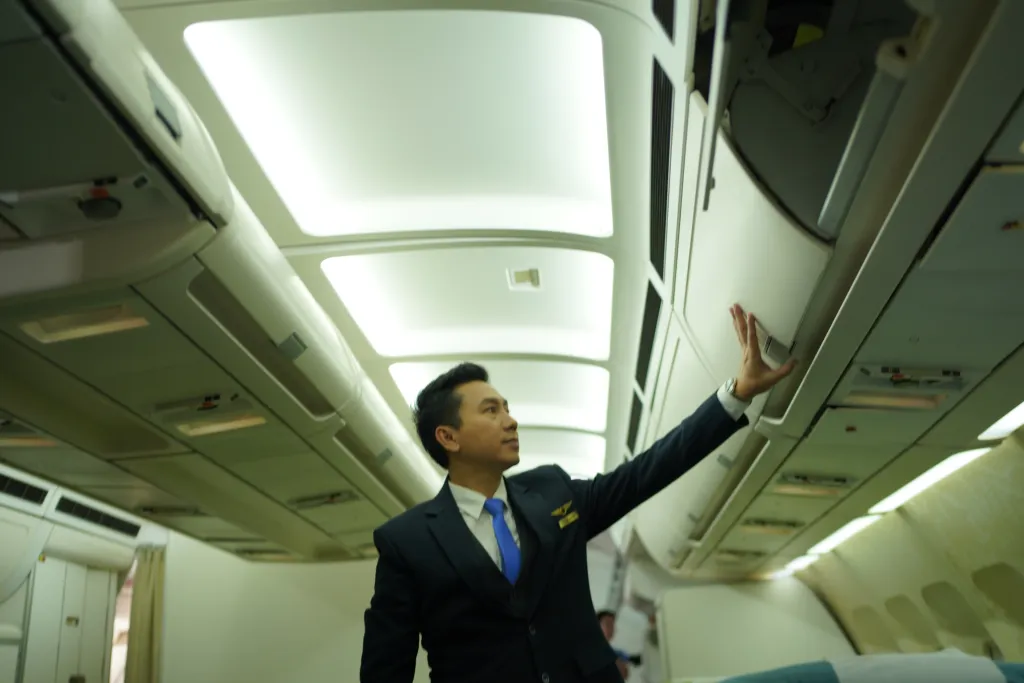 An Asian pilot in uniform stands by a passenger seat inside an airplane.