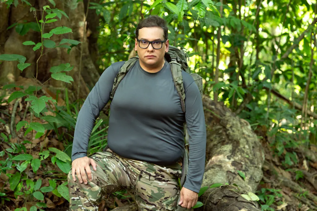George Santos in a jungle setting, wearing a long-sleeved gray shirt, camouflage pants, and a backpack.
