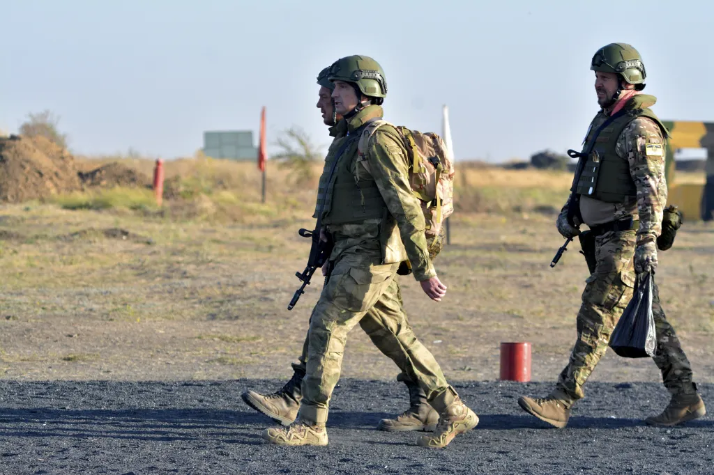 Two contract servicemen in camouflage uniforms and helmets walk across a dusty training ground, with one carrying a rifle and the other a black bag.