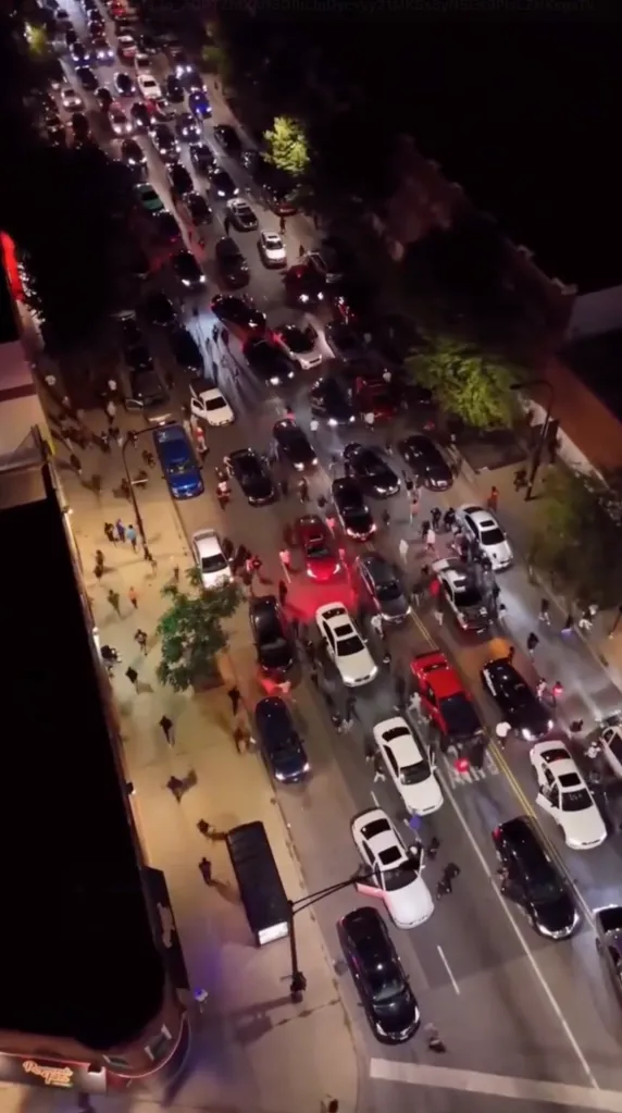 Aerial view of a street at night with many cars and people, some cars having doors open and red lights flashing.