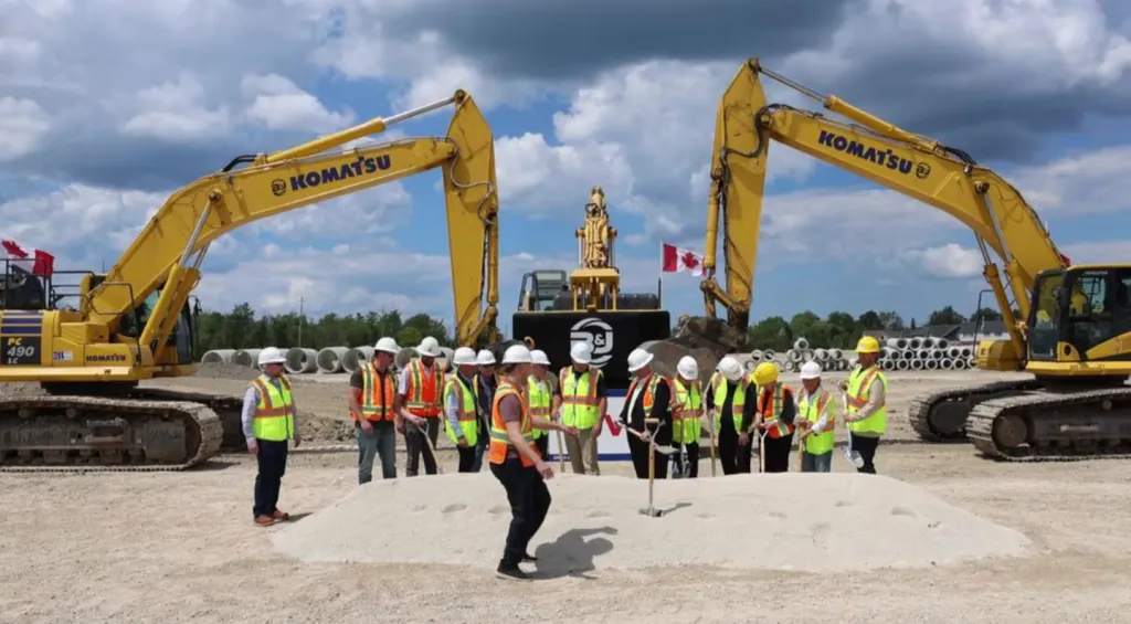 A group of construction workers in hard hats and safety vests breaking ground with shovels between two large yellow excavators.