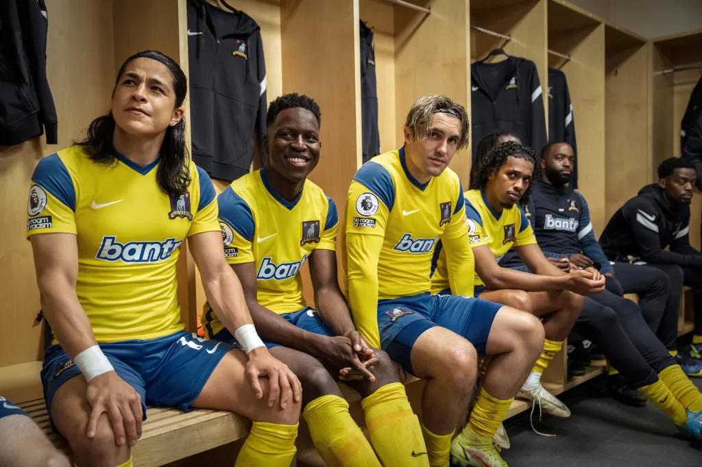 Ted Lasso actors Cristo Fernandez, Toheeb Jimoh, and Phil Dunster sit in a locker room in yellow and blue uniforms, with Moe Hashim and other teammates behind them.