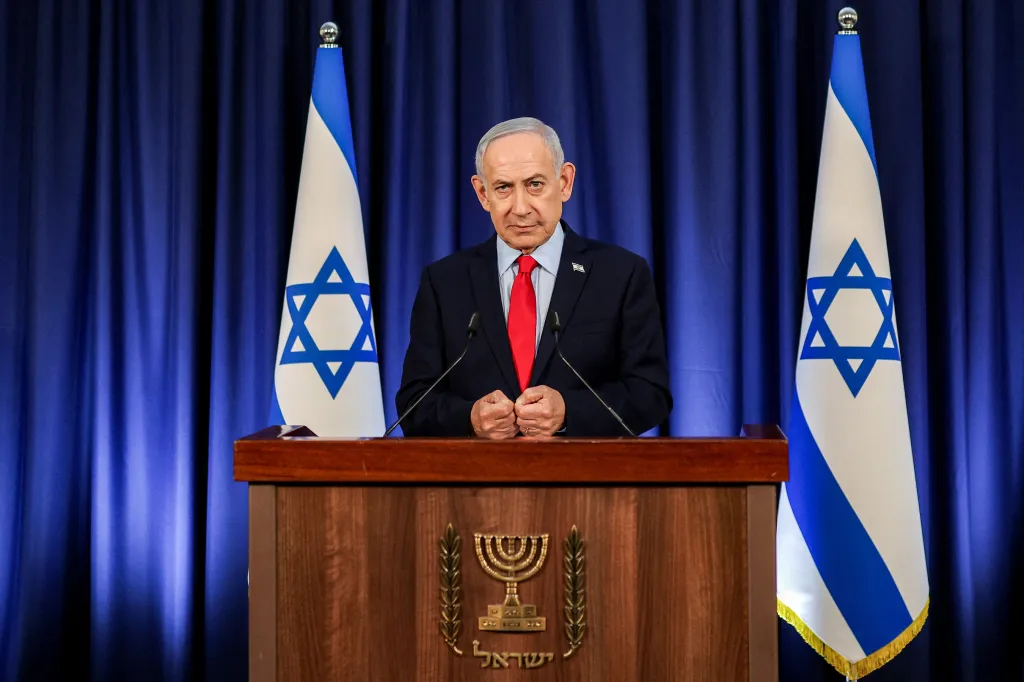 Israeli Prime Minister Benjamin Netanyahu speaks at a podium with two Israeli flags and a blue curtain in the background.