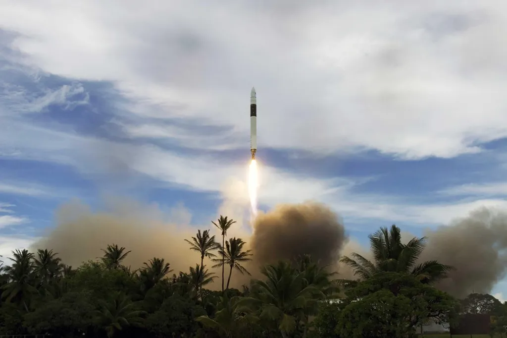 The Falcon 1 rocket launching with a fiery plume and thick smoke, seen rising above palm trees against a cloudy sky.