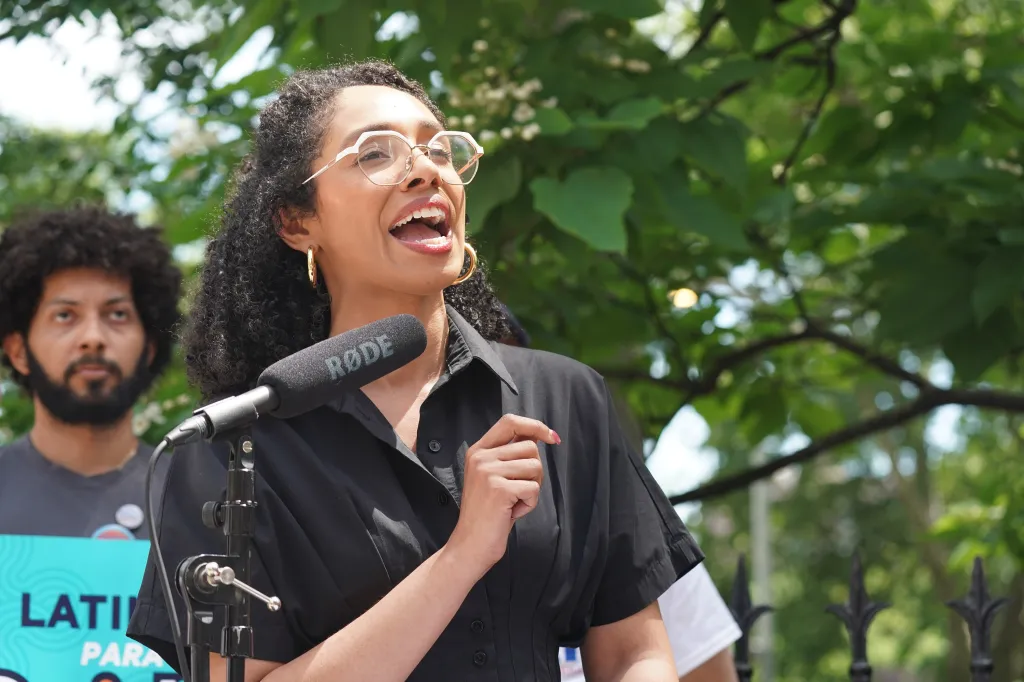 Darializa Avila Chevalier, wearing glasses and a black shirt, speaks into a microphone at an outdoor press conference.