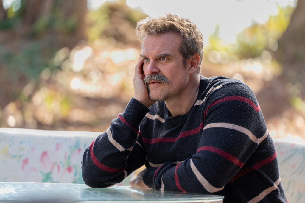 David Harbour with a mustache, wearing a striped sweater and resting his head on his hand.