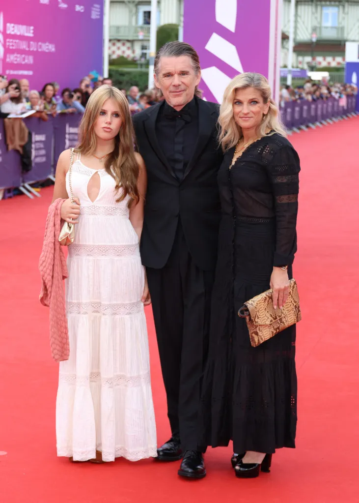 Ethan Hawke in an all-black suit with his daughter in a white dress and his wife in a black dress on a red carpet.