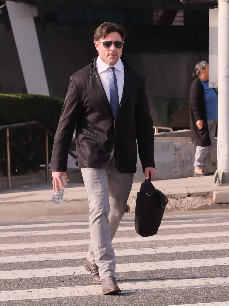 Aaron Phypers walks across a crosswalk carrying a water bottle and a briefcase.