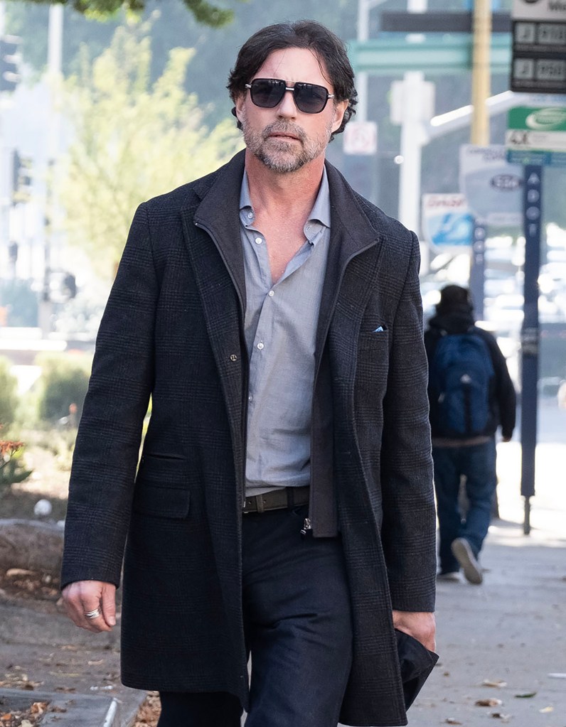Aaron Phypers walking outdoors wearing a long dark jacket, light gray shirt, and dark pants.