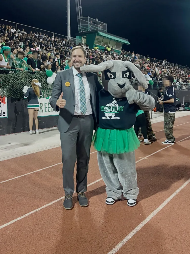 Derek Marshall, candidate for Victorville City Council, standing next to a person in a gray rabbit mascot costume, giving a thumbs up at a football game.