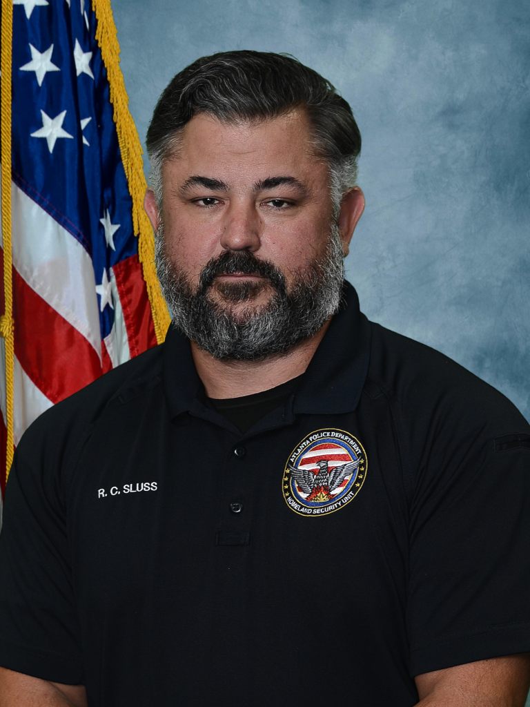 Headshot of Detective Ronald Sluss wearing a black polo shirt with an Atlanta Police Department patch and an American flag in the background.