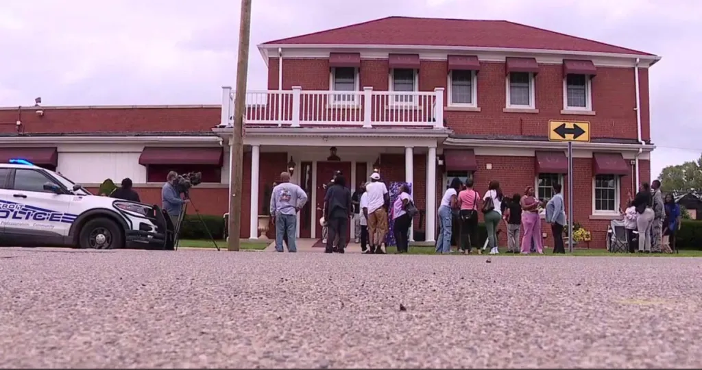 A crowd of people, including a cameraman and a police car, outside a red brick building with red awnings and a balcony.