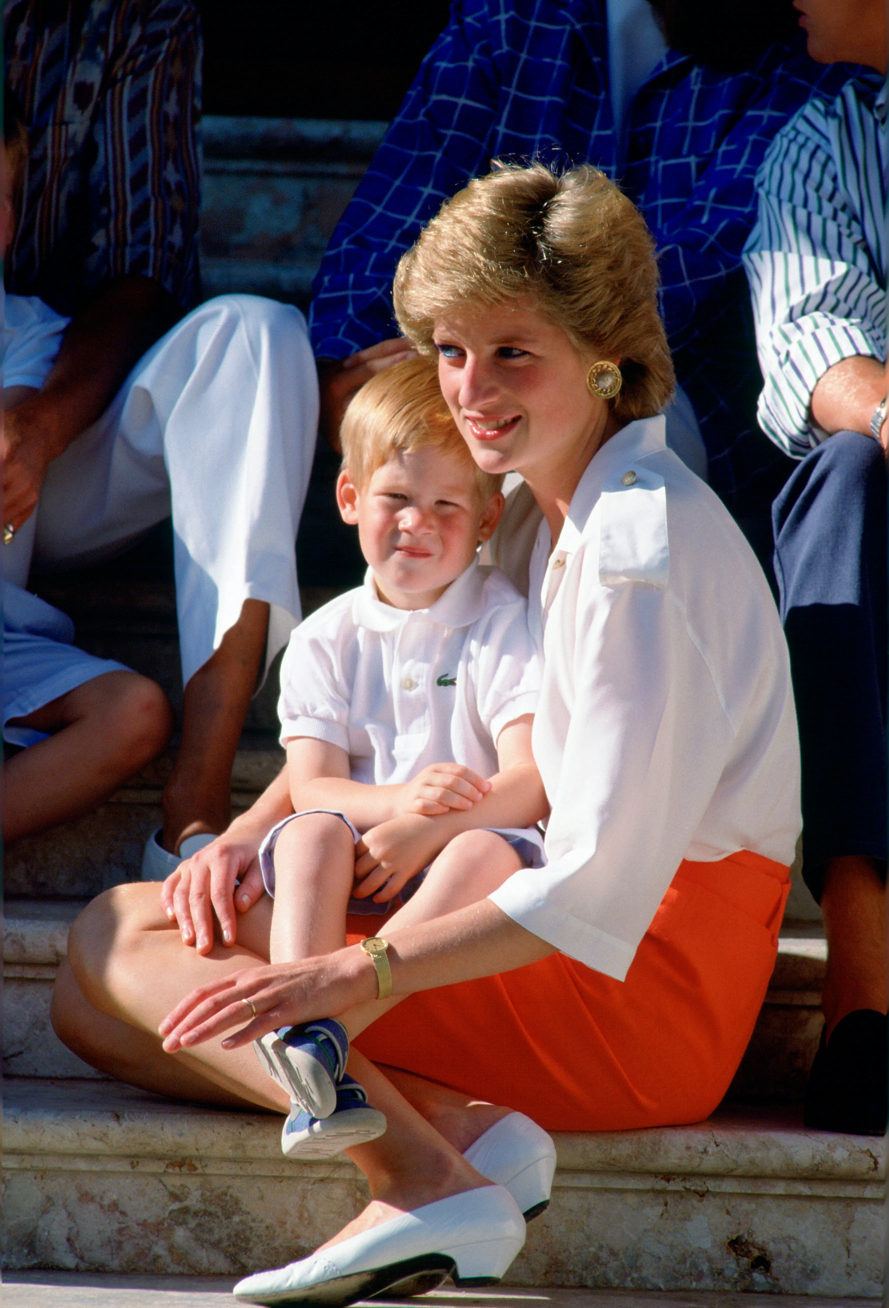Princess Diana in a white blouse and orange skirt, holding young Prince Harry in a white Lacoste shirt on her lap, sitting on steps.