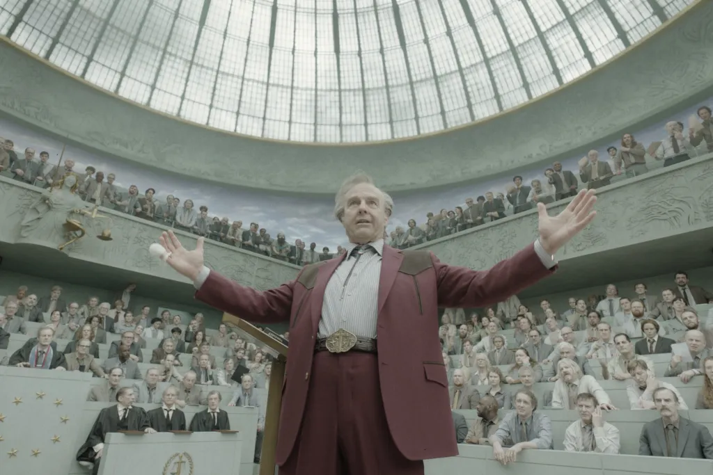 Tom Cruise, wearing a burgundy suit, before an audience in a domed hall.