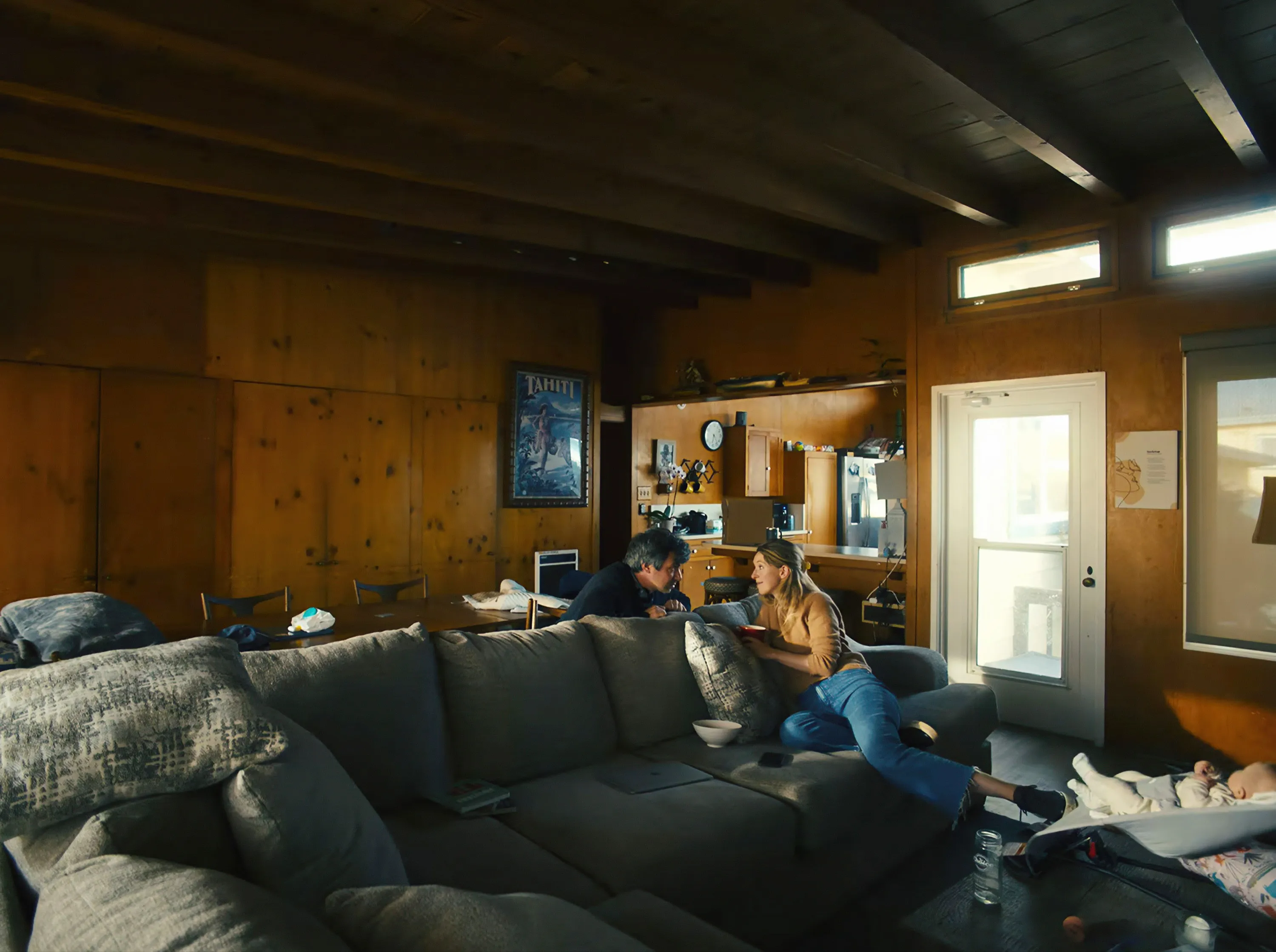 Nathan Fielder and Elizabeth Holmes talking on a couch with a baby in a bassinet nearby.