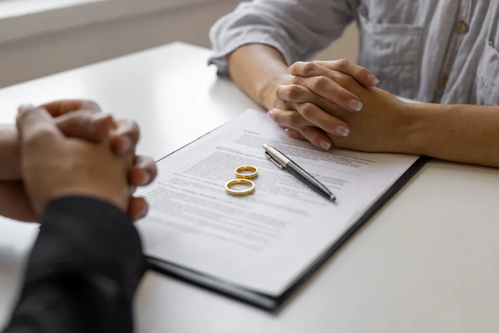 Two people sitting at a table with a divorce agreement, pen, and two wedding rings.
