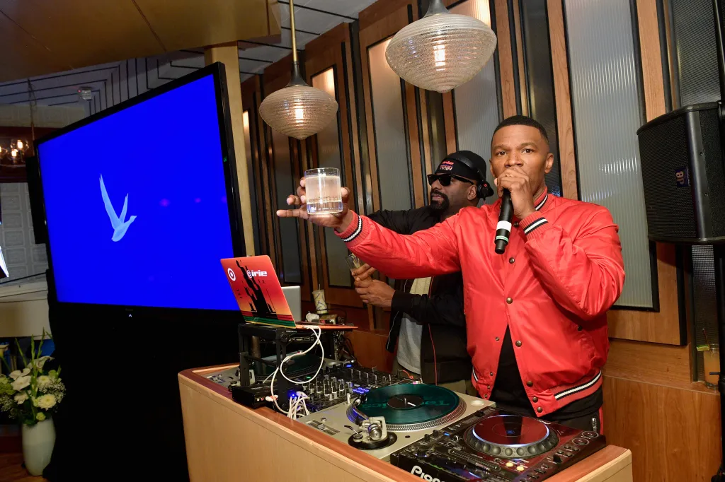 Jamie Foxx at a DJ booth, holding a microphone in one hand and a drink in the other, with DJ Irie behind him.