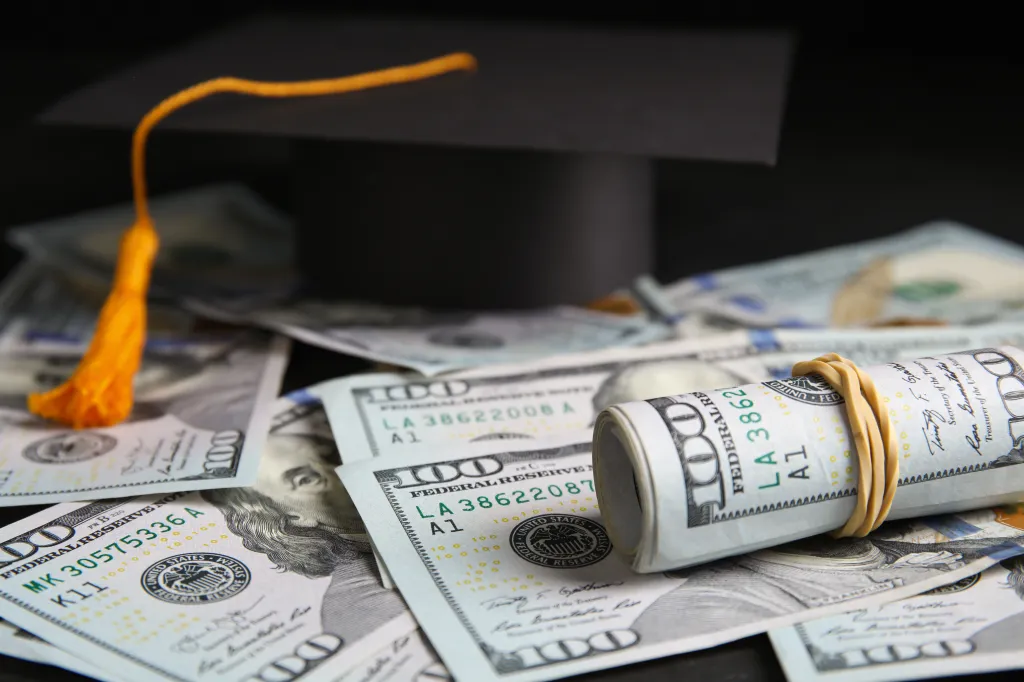 A graduation cap rests on a scattered pile of US hundred-dollar bills, with one stack of bills rolled and secured with a rubber band.