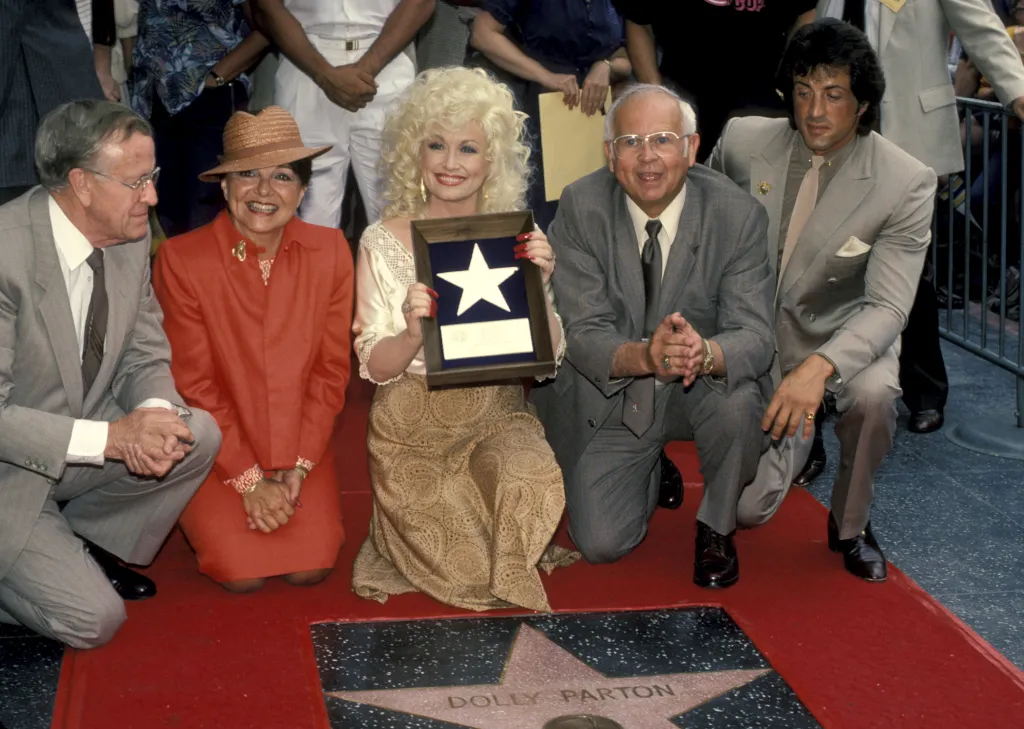 Dolly Parton, holding a framed star award, kneeling with her family and Sylvester Stallone on the Hollywood Walk of Fame.