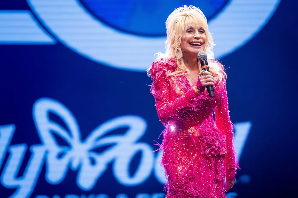 Dolly Parton in a pink dress, holding a microphone and smiling on stage at Dollywood.