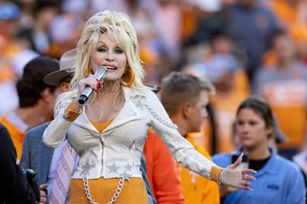 Dolly Parton performs in a sparkly white jacket with orange accents, holding a microphone.