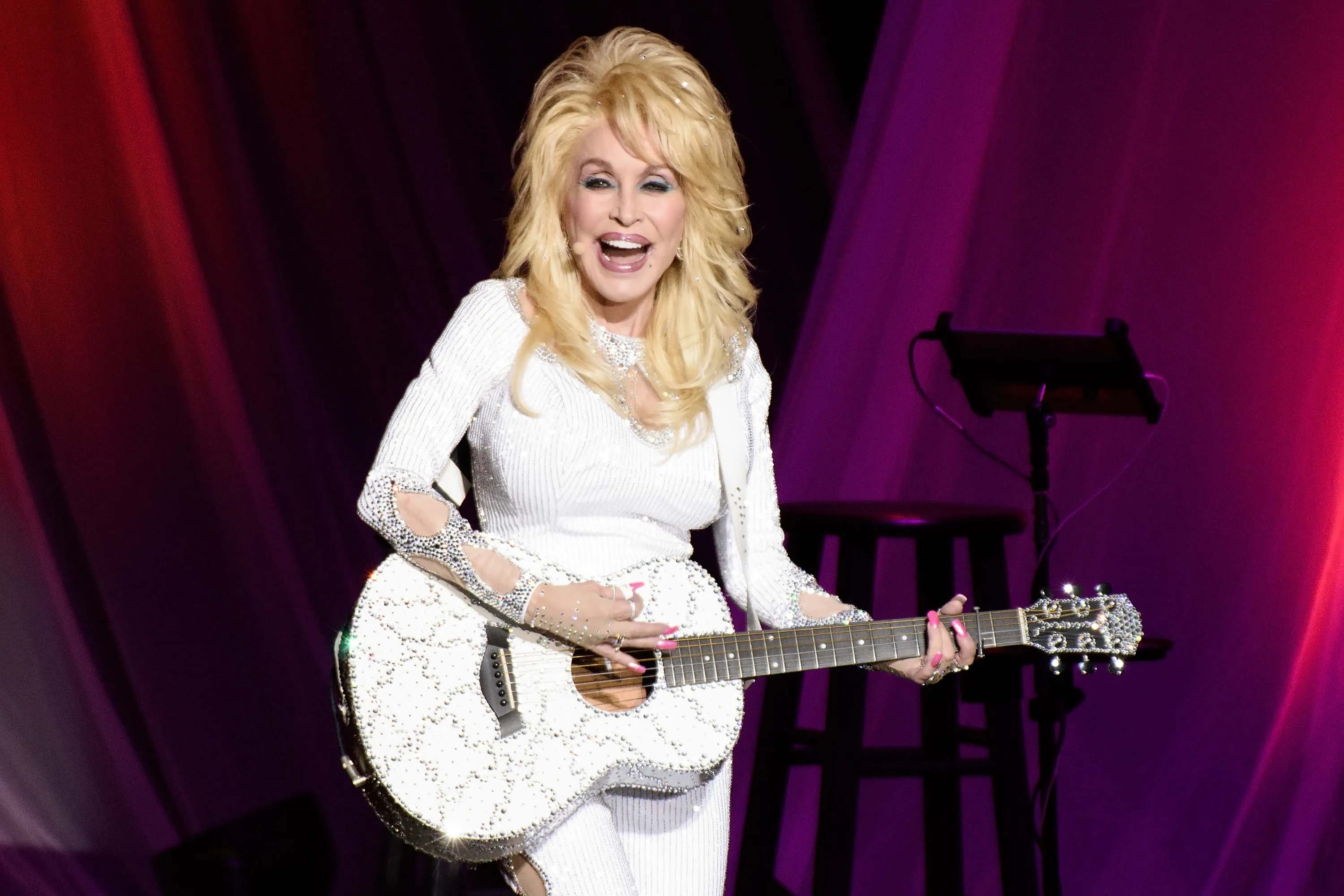 Dolly Parton performing in a white, sparkly outfit while playing a matching sparkly white guitar.