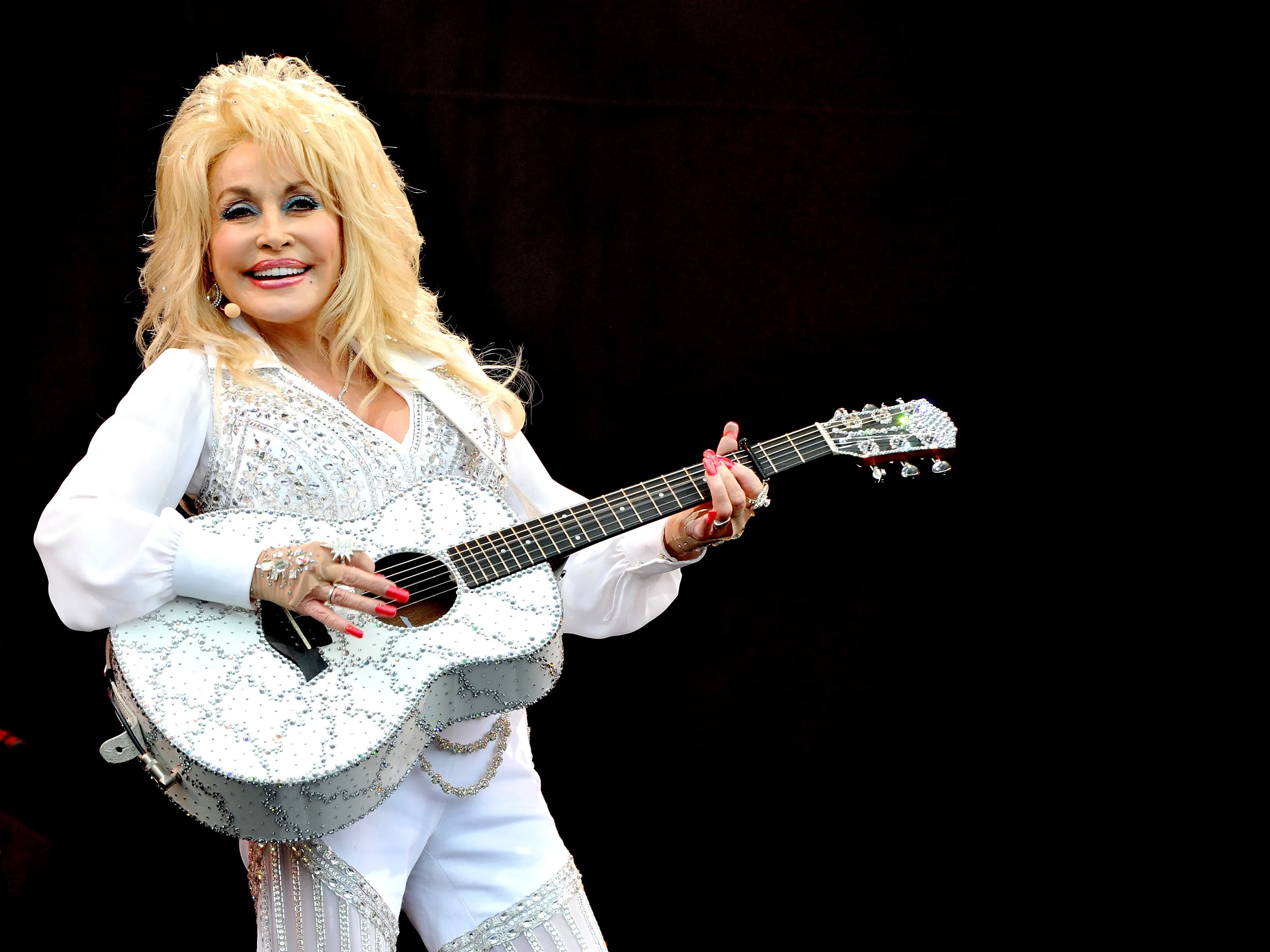 Dolly Parton in a white rhinestone outfit and matching guitar performing on stage.