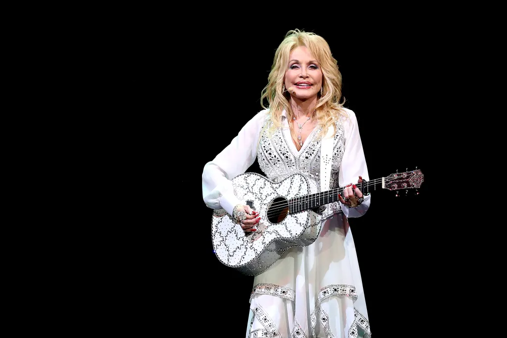 Dolly Parton performs on stage, playing a white bejeweled guitar.