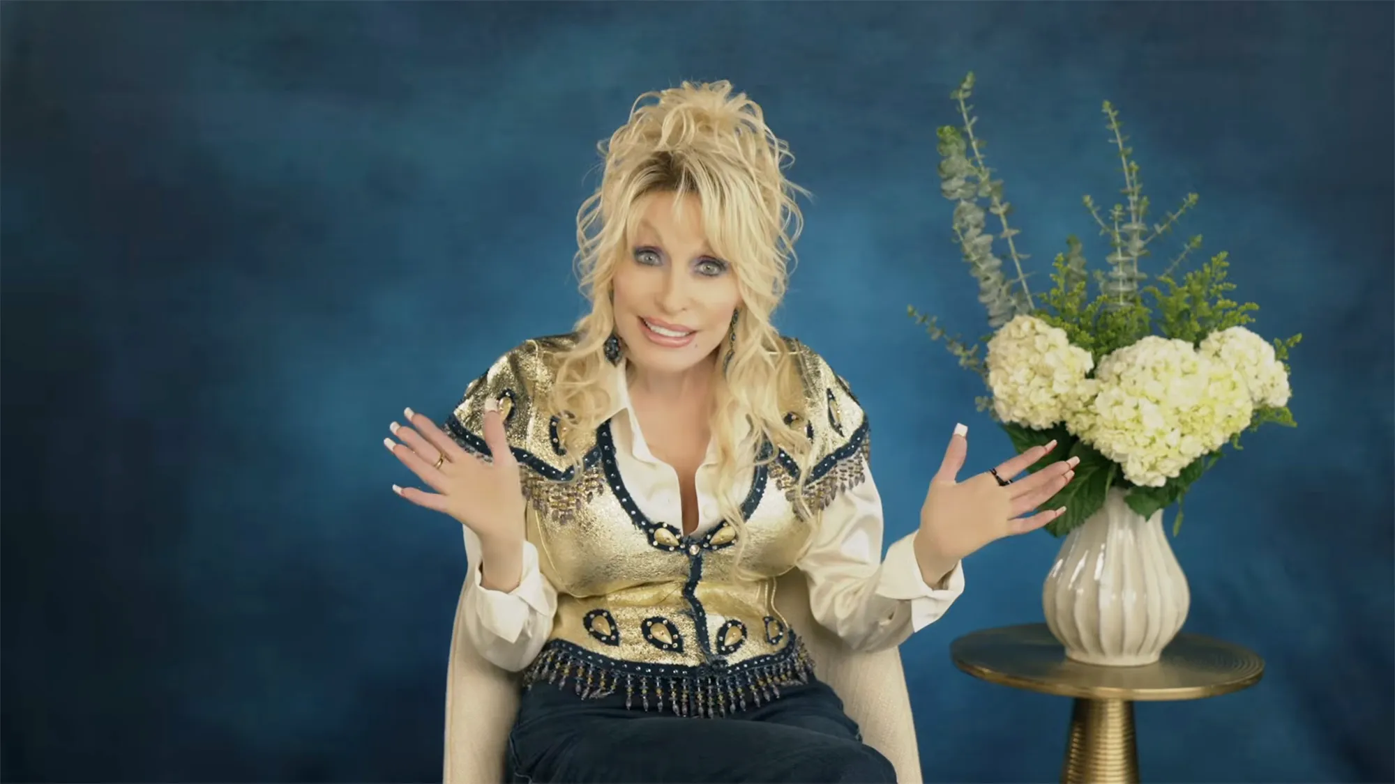 Dolly Parton wearing a gold and blue embellished top, with her hands open and a bouquet of flowers next to her.