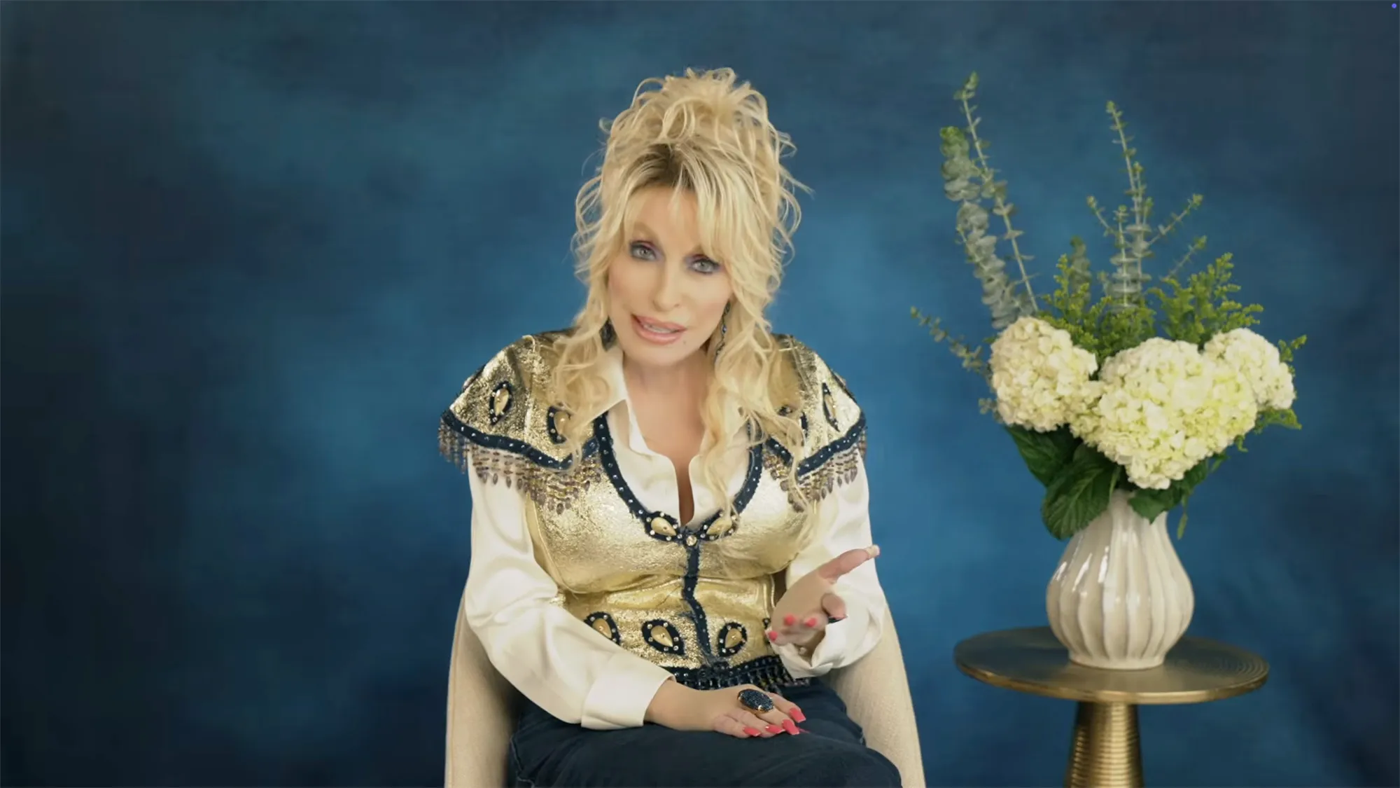 Dolly Parton speaking while wearing a gold vest and white shirt.