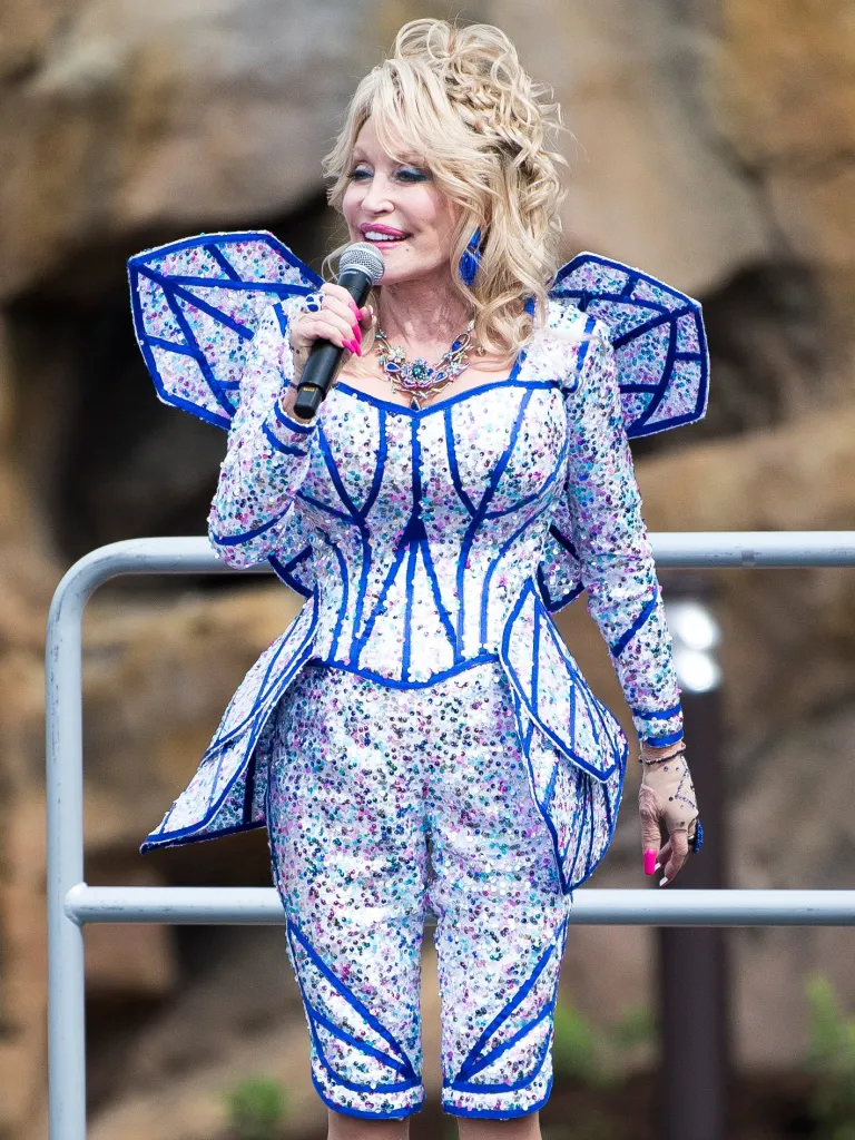 Dolly Parton in a sparkly white and blue outfit speaks into a microphone at Dollywood's Wildwood Grove expansion.