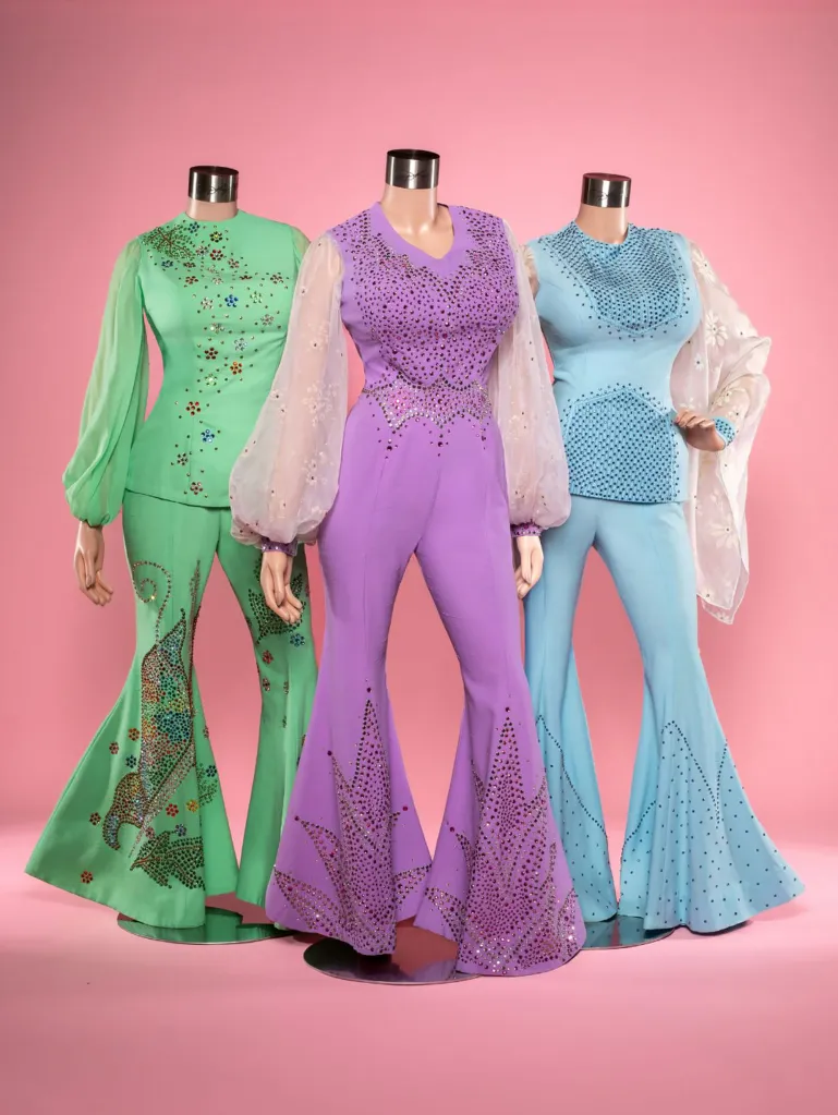 Three mannequins display Dolly Parton's outfits featuring embellished flared pants in green, purple, and blue against a pink background.