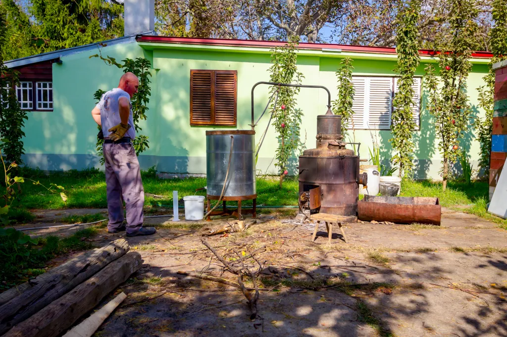 A man stands next to a homemade copper distillery, making alcohol outside a light green building.