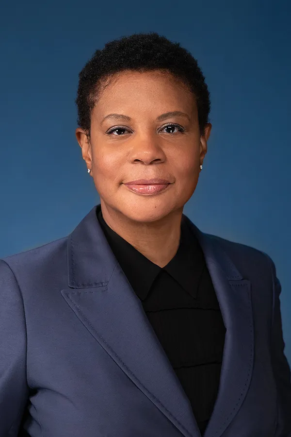 Dr. Alondra Nelson smiling, wearing a dark suit jacket and black shirt against a blue background.