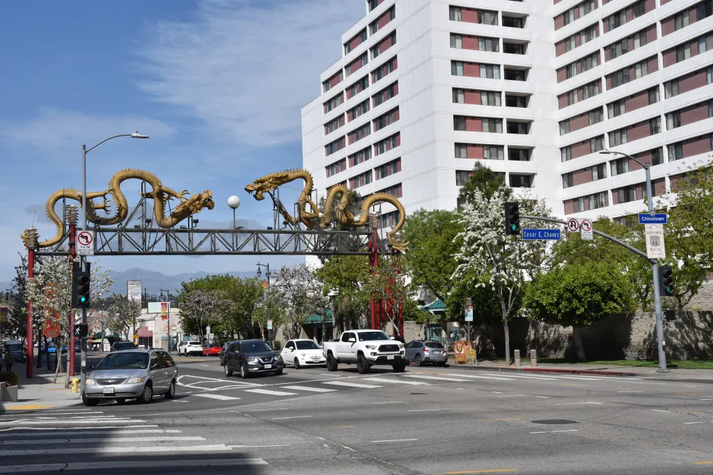 The Dragon Gate, featuring two golden dragons, marks the entrance to Los Angeles Chinatown, with a white building and street scene in the background.