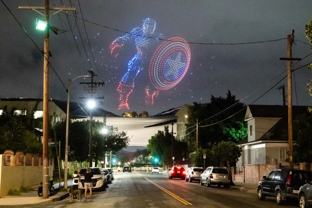 A drone show forms the shape of Captain America, red and blue against a dark sky, above a street in Los Angeles.
