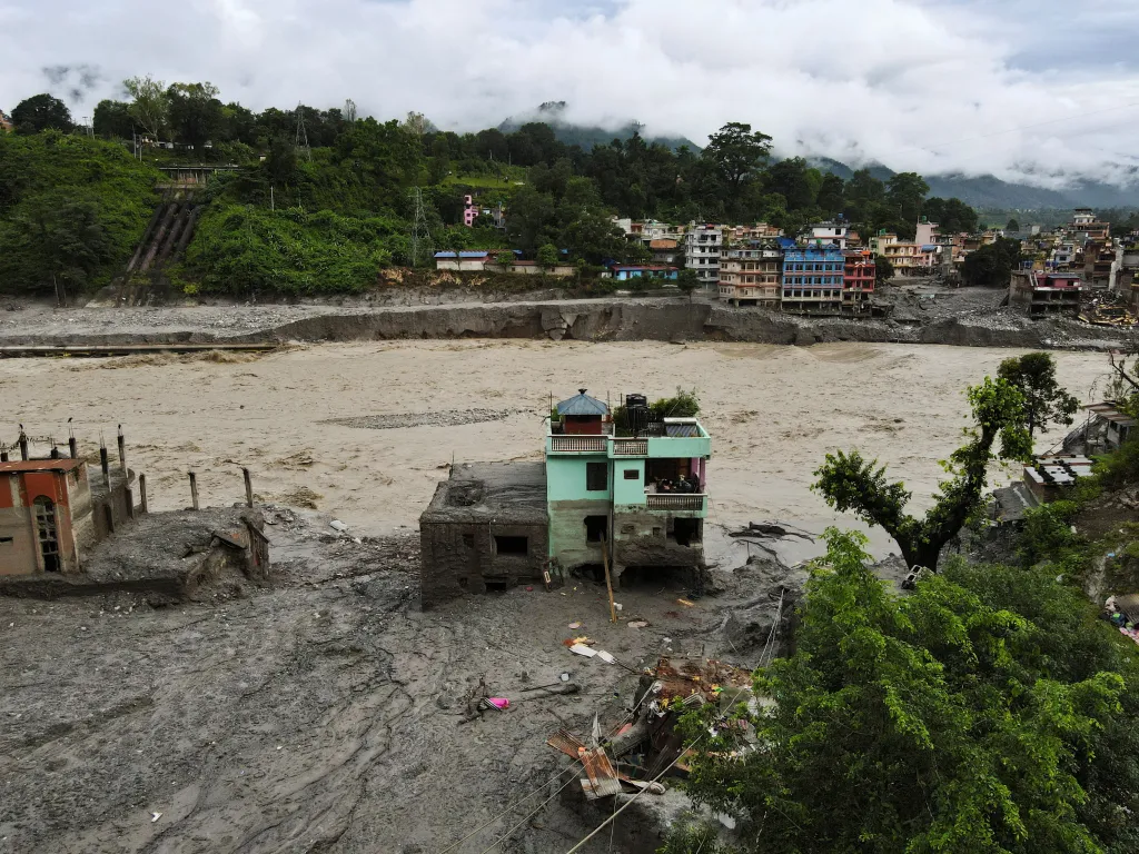 Drone view of a light green multi-story house standing amid mud and debris next to a wide, muddy river, with buildings and green hills in the background.