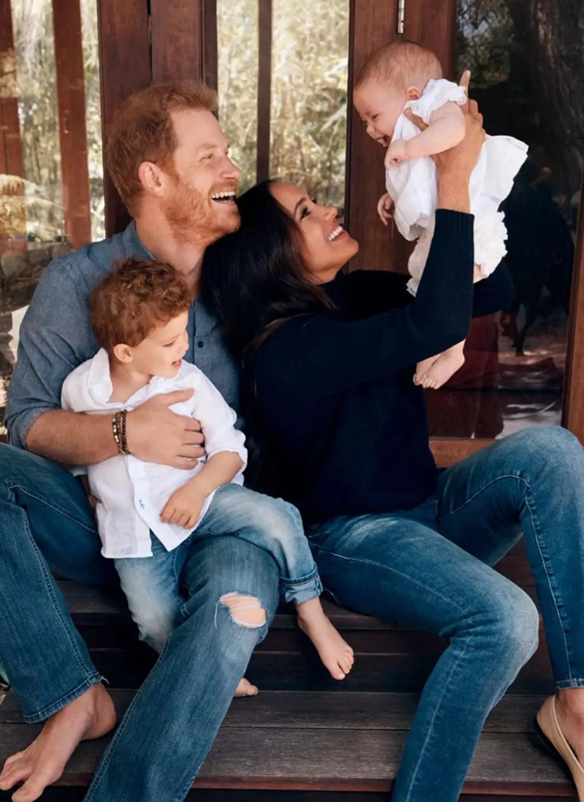 The Duke and Duchess of Sussex smiling and looking at their children, Meghan holding their daughter Lilibet in the air, and Prince Harry holding their son Archie on his lap.