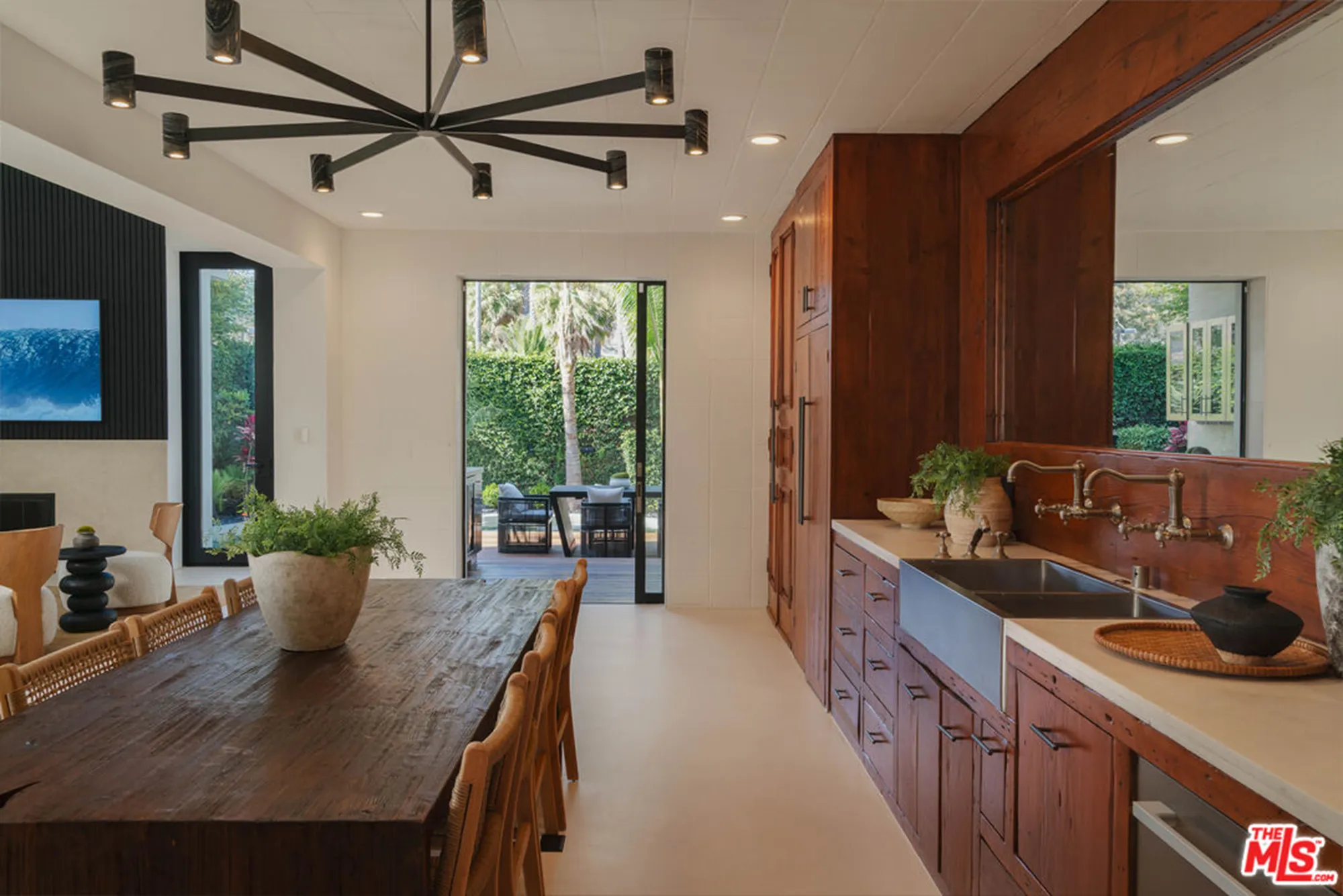 Dining area and kitchen of a home, with a long wooden table, modern lighting, and an outdoor patio visible through open doors.