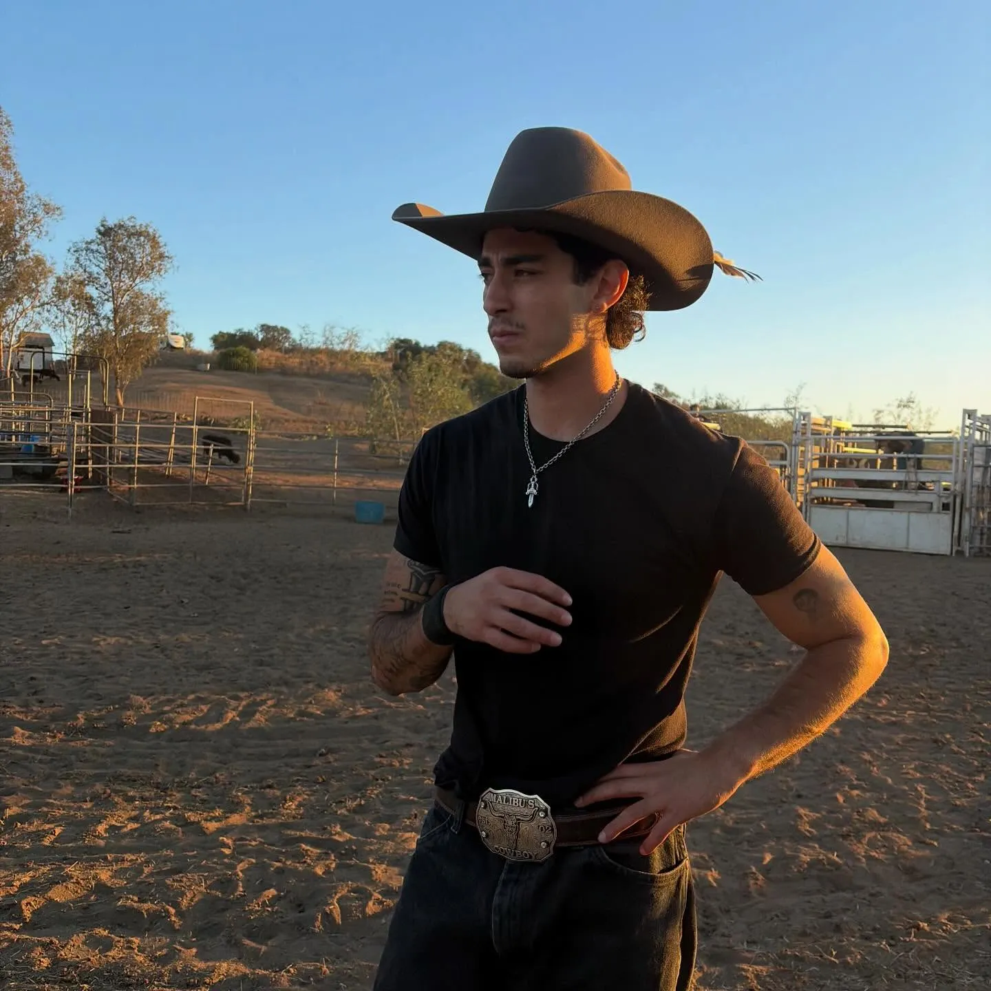 A young man in a cowboy hat and black t-shirt standing in a fenced dirt arena at sunset.