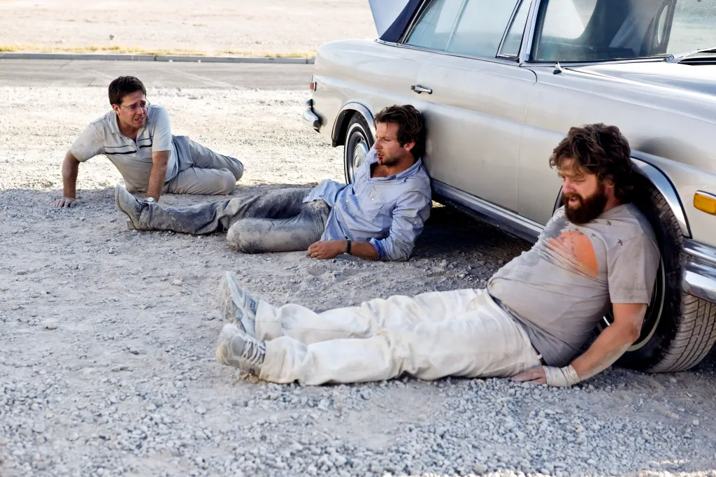 Ed Helms, Bradley Cooper, and Zach Galifianakis lying on the ground next to a car in a scene from the film 