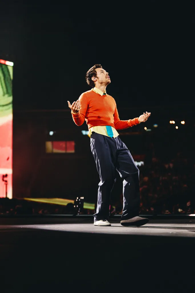 Harry Styles performing on stage, wearing an orange sweater, blue trousers, and a yellow shirt with a blue tie, looking up and gesturing with both hands.