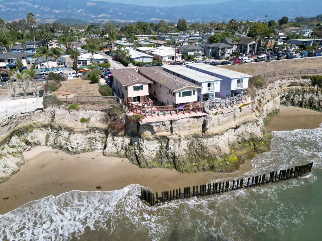 Aerial view of Isla Vista residences sitting on a cliff threatened by collapse, with the ocean waves crashing onto the sandy beach below.