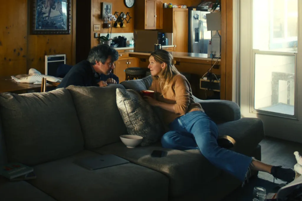 Elizabeth Holmes, founder and former CEO of Theranos, sits on a couch in conversation with Nathan Fielder in her home.