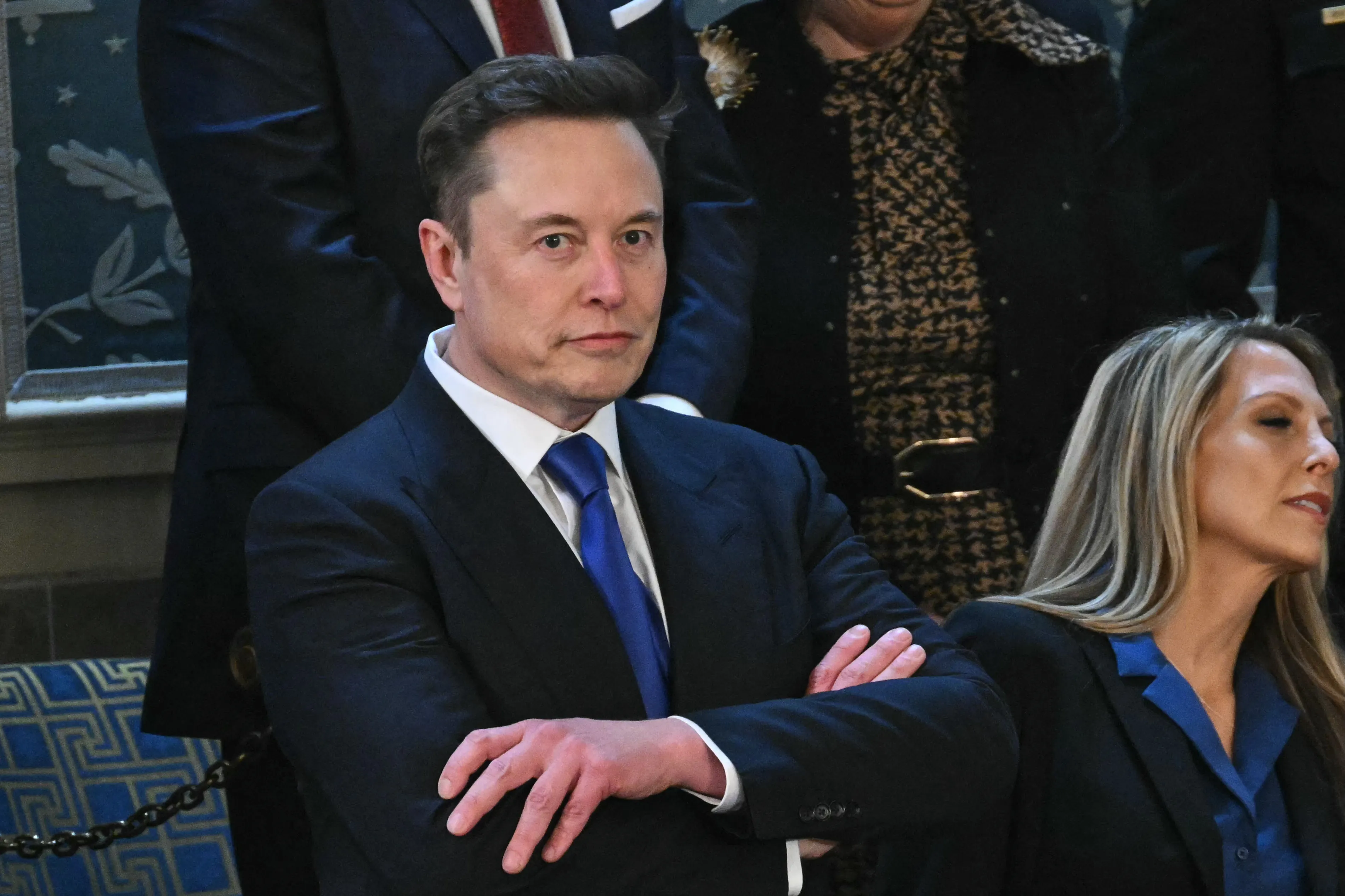 Elon Musk in a dark suit and blue tie, seated with his arms crossed, attending President Trump's address.