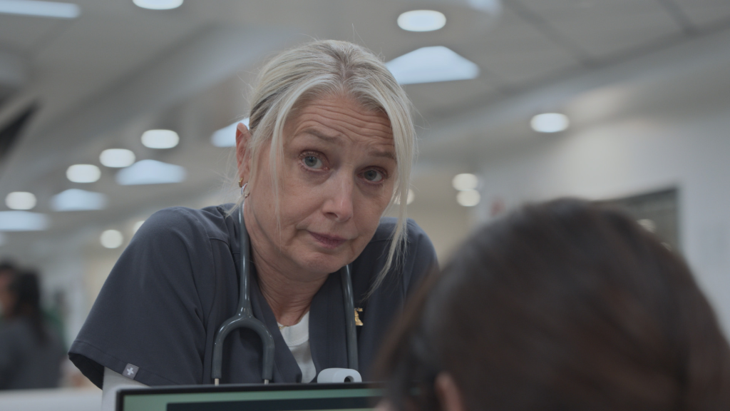 Katherine LaNasa as Nurse Sue approaching someone from behind a desk.