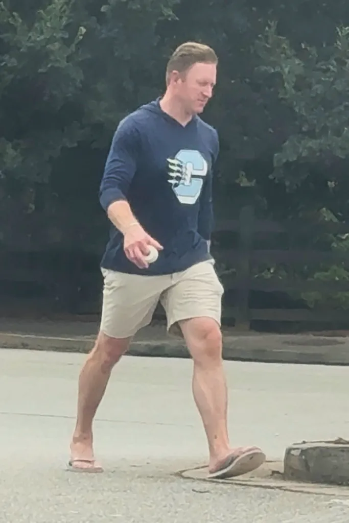 Kroy Biermann in a navy hoodie and beige shorts, holding a white baseball, walking away from his son's game.