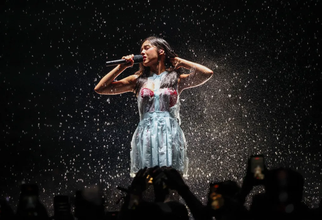 Olivia Rodrigo performing on stage in a light blue dress with red details, wet from water spraying around her.