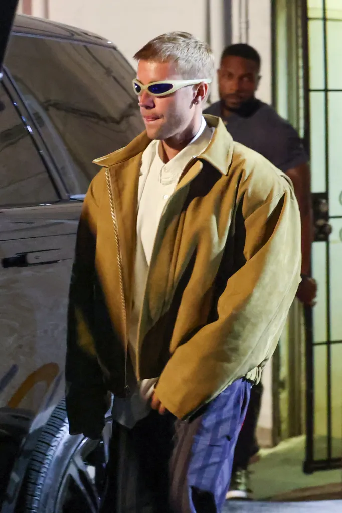 Justin Bieber in a brown jacket, white polo shirt, blue pants, and white-framed sunglasses.