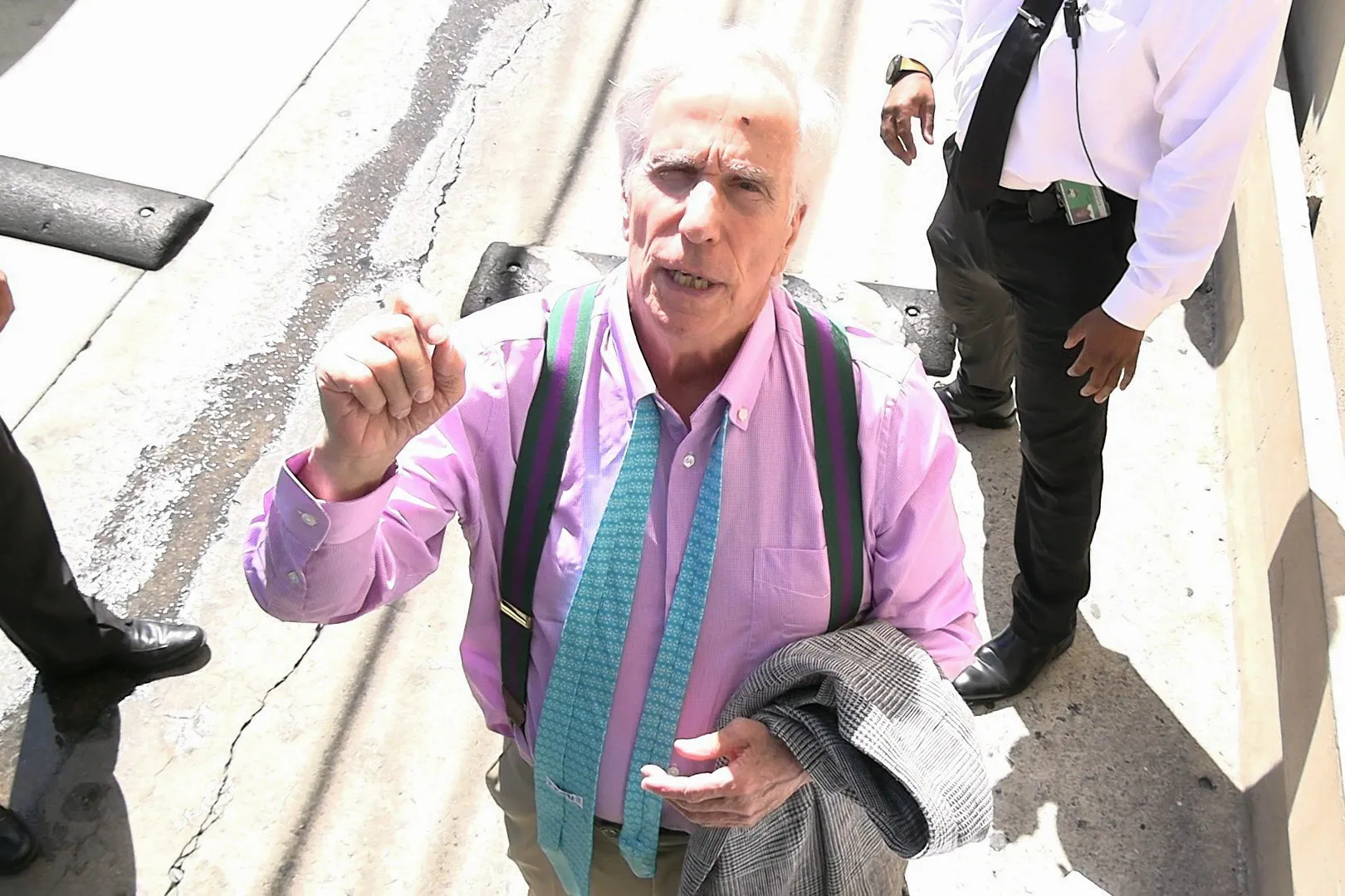 Henry Winkler in a pink shirt, green and purple suspenders, and a turquoise tie talks to photographers.